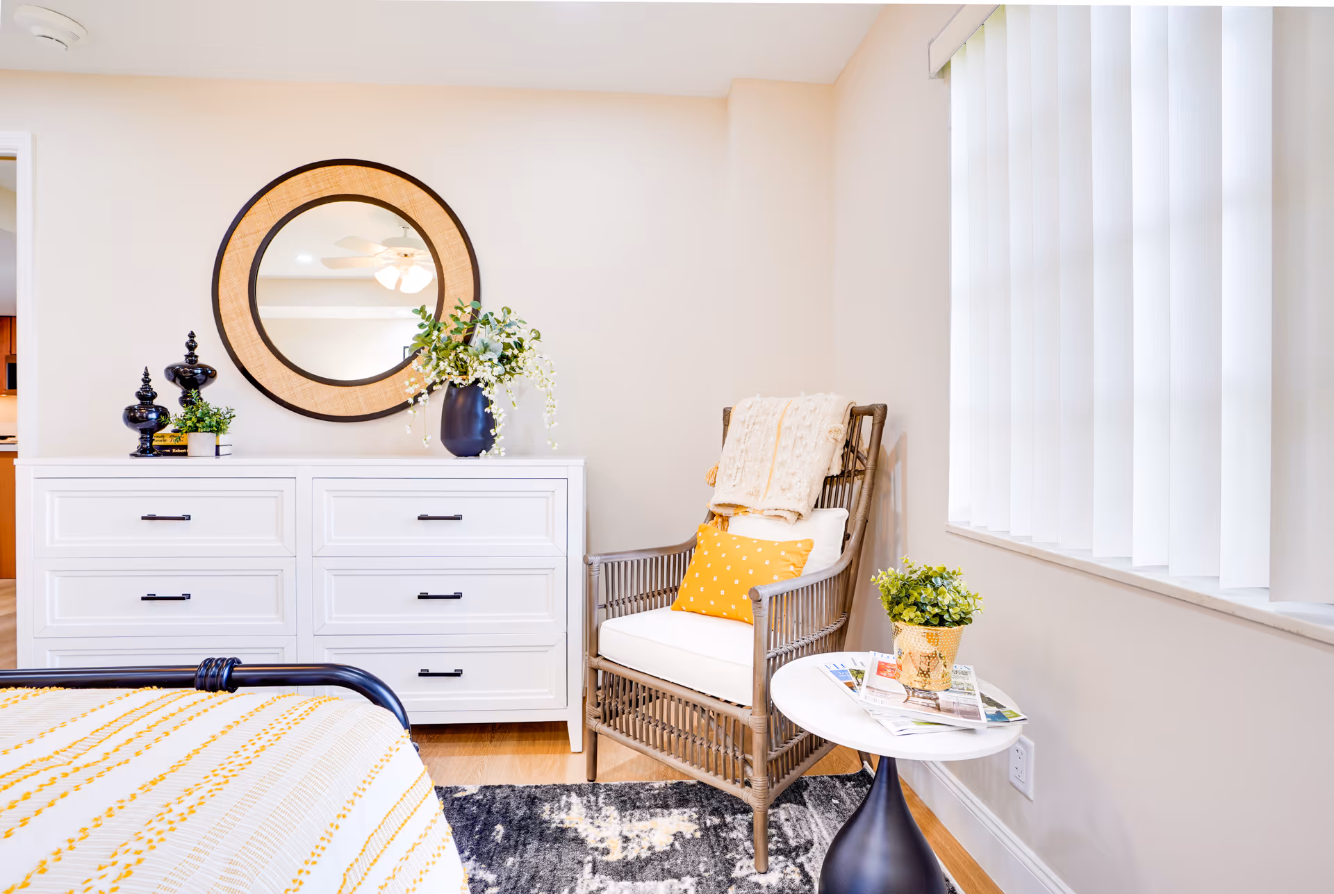 A cozy corner of a bedroom featuring a white dresser with black handles, a round mirror with a woven frame above it, and decorative items including a black vase with white flowers and small black ornaments. Next to the dresser is a wicker armchair with a white cushion, a yellow polka dot pillow, and a beige throw blanket. A small round white table with a black base holds a potted plant and some magazines. Vertical blinds cover the window on the right side.