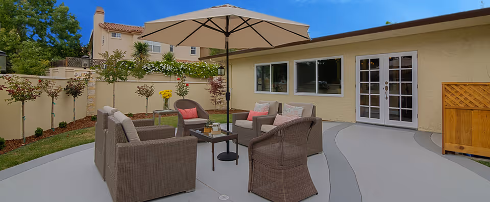 Outdoor patio area with wicker furniture including two armchairs, a loveseat, and a coffee table under a large beige umbrella. The patio is surrounded by a low wall with small trees and plants along the edge. A building with beige walls, windows, and French doors is visible in the background.