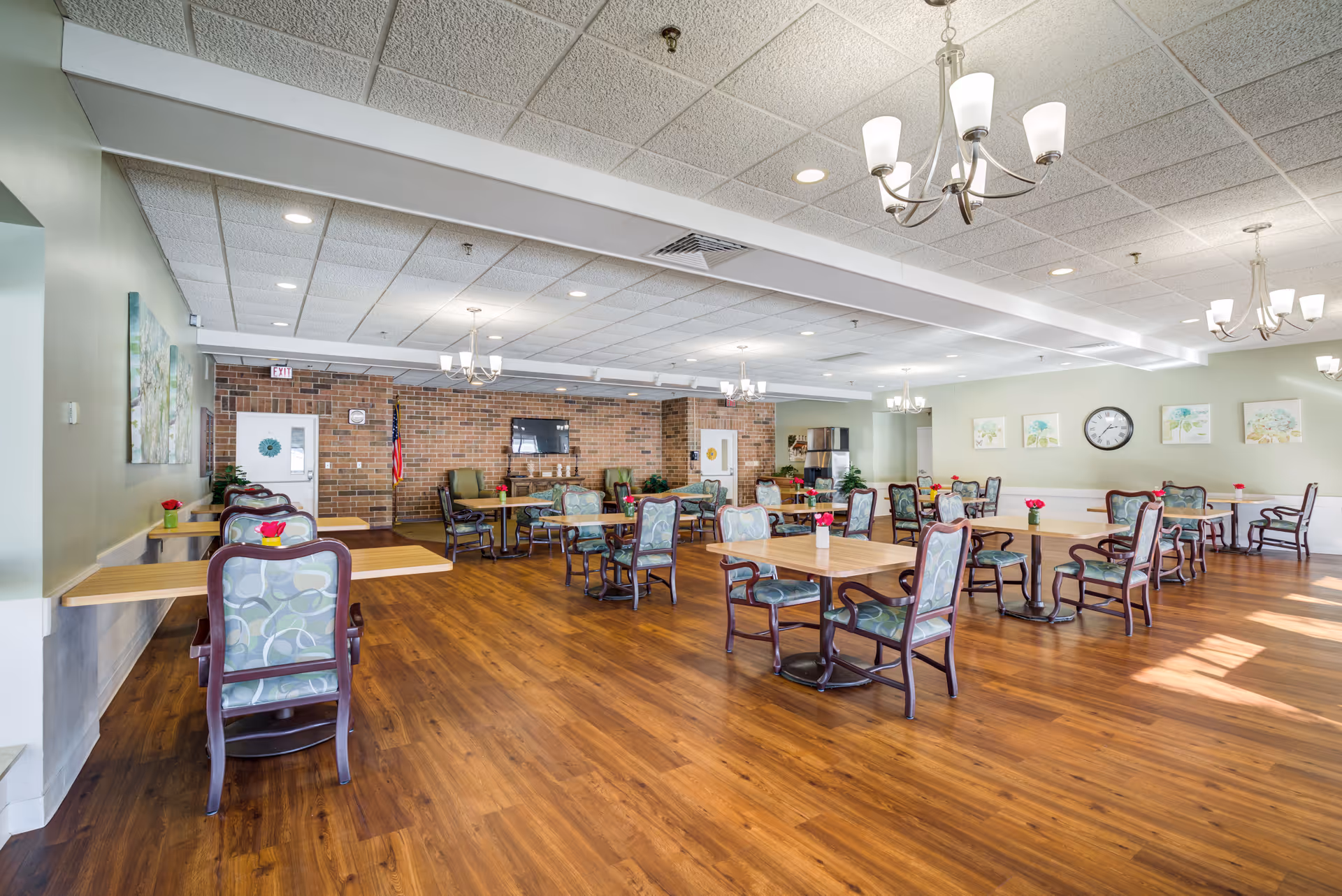 A spacious dining room with wooden floors, multiple tables and chairs with patterned upholstery, chandeliers hanging from a white tiled ceiling, a brick accent wall with a mounted TV, an American flag, and several framed artworks on the light green walls.