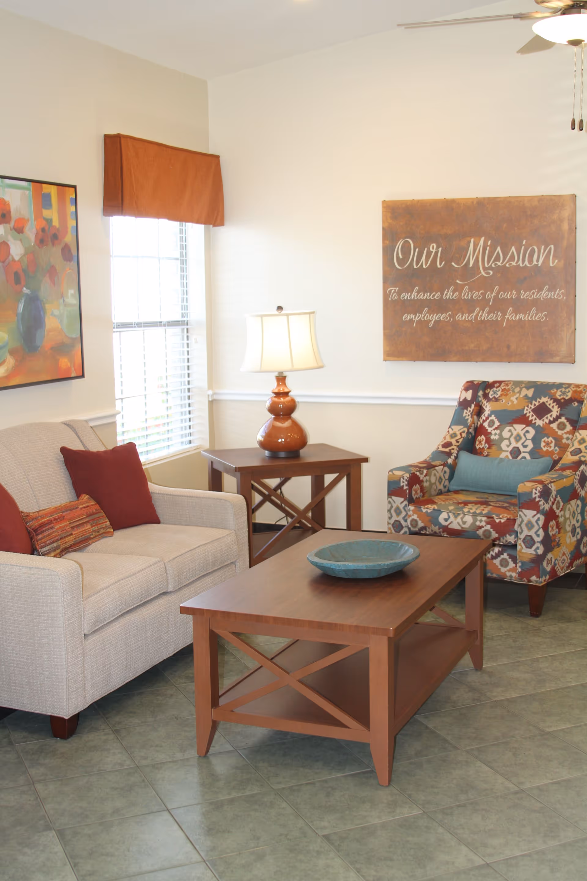 A bright sitting area with a sofa, patterned armchair, wooden coffee and side tables, a lamp, and wall art that reads "Our Mission".