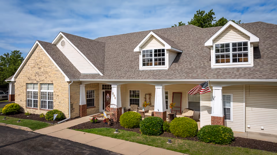 Exterior view of a single-story assisted living facility building with beige siding and stone accents, featuring a covered porch with chairs and an American flag, surrounded by neatly trimmed bushes and a paved walkway.