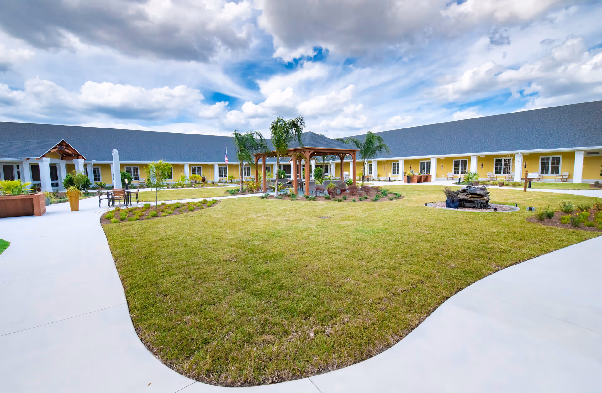 Well-maintained courtyard with a central gazebo, lawn, walkways, fountain and a surrounding single-story yellow building under a partly cloudy sky.