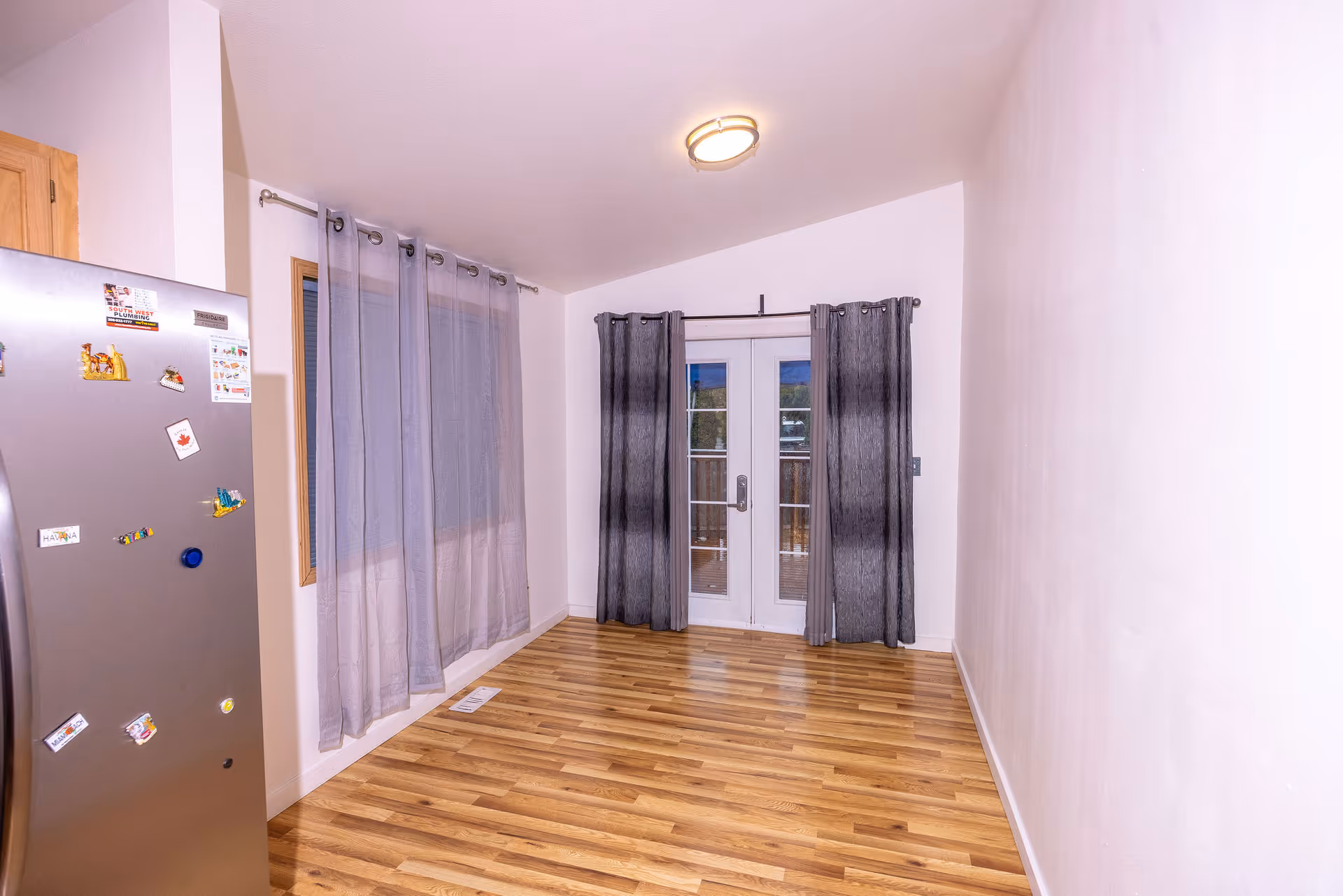 Empty room with wooden floor, white walls, a stainless steel refrigerator with magnets on the left, two windows with sheer gray curtains, and a set of French doors with dark gray curtains leading outside.