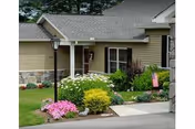 Exterior view of a single-story residential building with beige siding and a gray roof. The front yard features a well-maintained garden with various colorful flowers and shrubs, a lamp post near the walkway, and a small porch area with a white column.