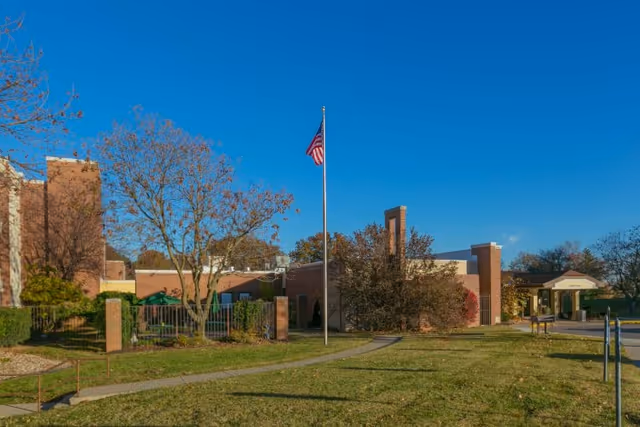 Exterior view of a brick building with a flagpole flying the American flag in front, surrounded by trees and a grassy lawn under a clear blue sky.