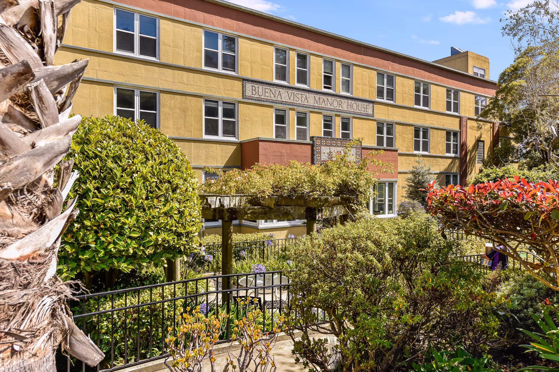 Exterior view of Buena Vista Manor House, a multi-story building with yellow and reddish-brown walls, surrounded by lush greenery, bushes, and trees under a blue sky with some clouds.