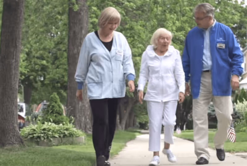 An elderly woman walking outdoors on a sidewalk accompanied by two caregivers, one on each side, with trees and greenery in the background.