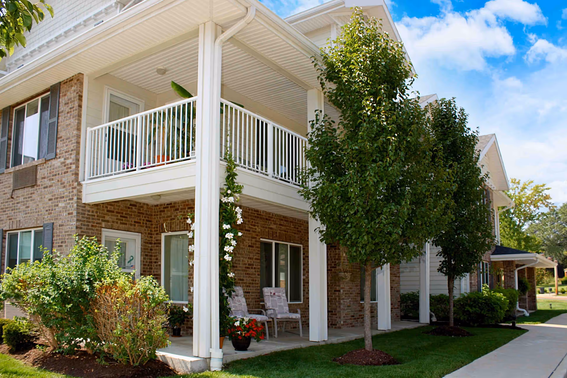 Exterior view of a two-story brick and siding building with a covered porch and balcony. The porch has two cushioned chairs and potted plants, and there are small trees and shrubs planted along the walkway. The sky is partly cloudy and the area looks well-maintained.
