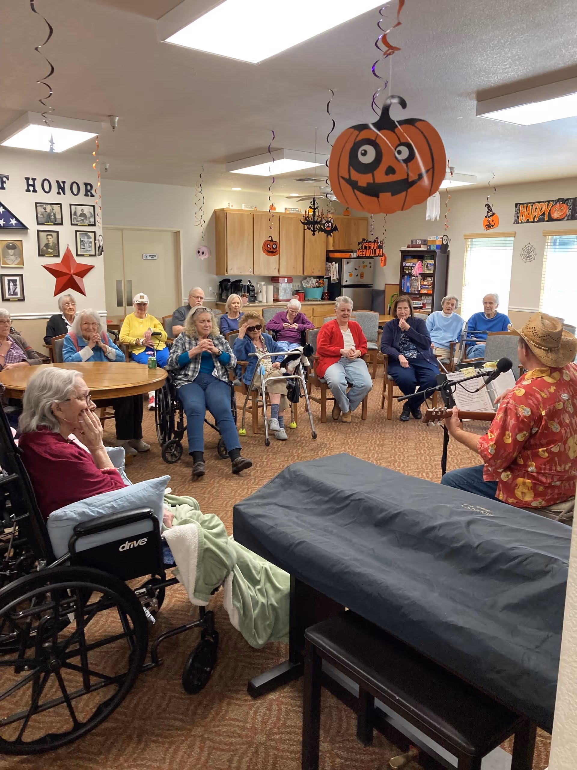 A group of elderly people seated in a common room decorated with Halloween-themed hanging decorations, including pumpkins and spiders. They are attentively watching a man wearing a cowboy hat and a festive shirt playing a guitar and singing into a microphone. The room has a piano covered with a black cloth and a wall display with photos and a red star.