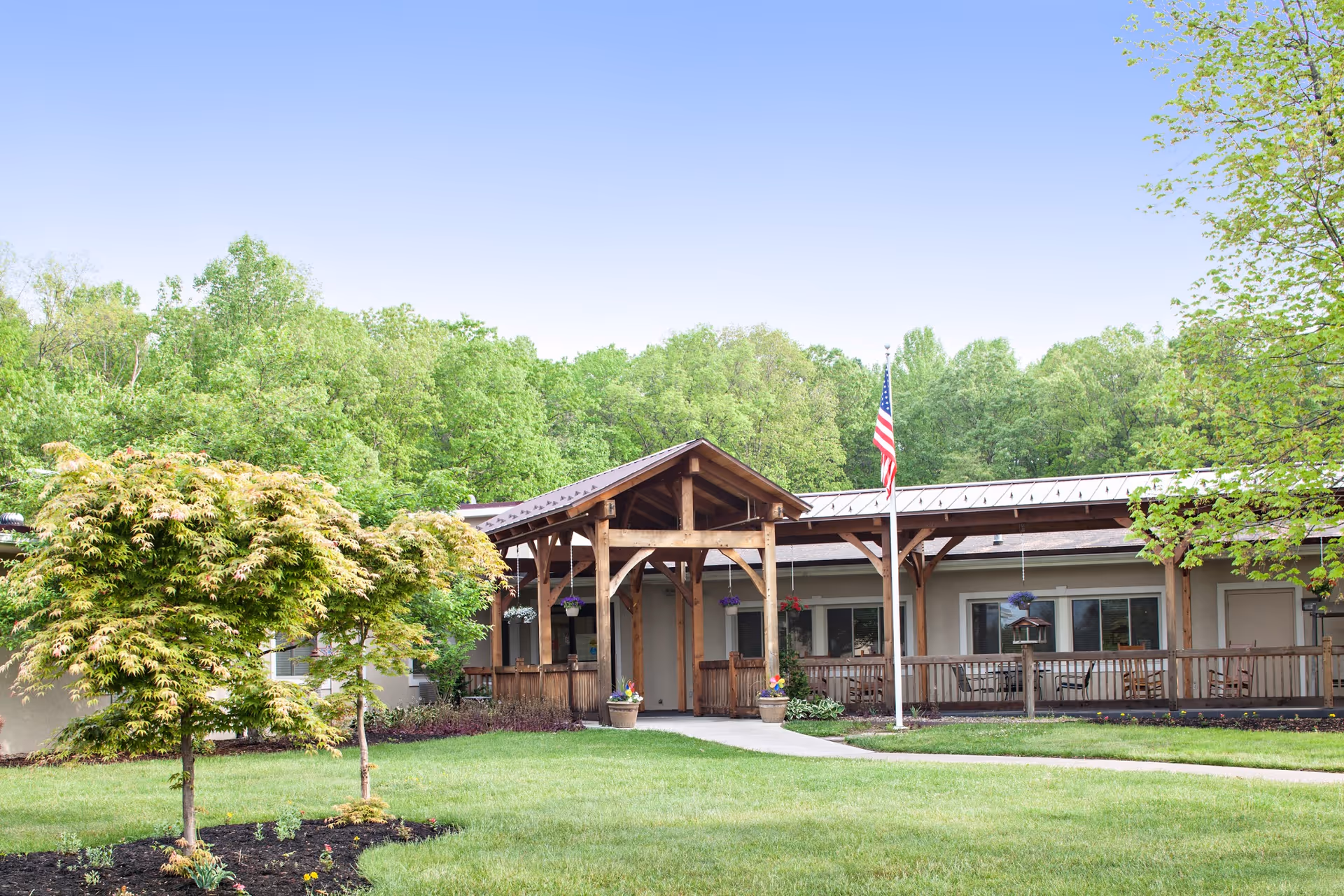 Front exterior of a single-story senior living building with a covered wooden entrance, American flag, and landscaped lawn and trees.