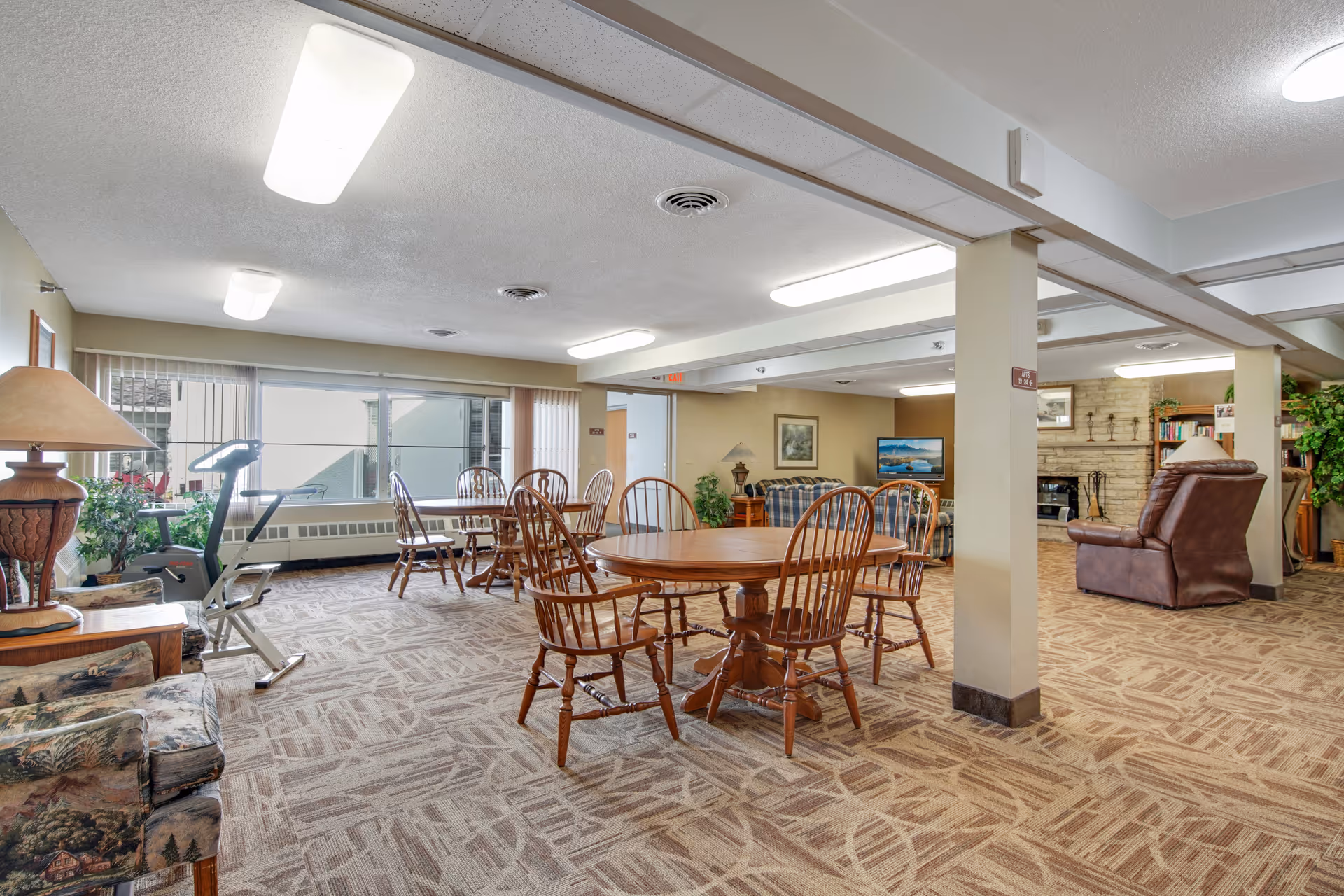 A spacious common area in an assisted living facility featuring multiple wooden tables with chairs, a seating area with armchairs and a couch, a television, a fireplace, and large windows letting in natural light. There is also an exercise bike near the window and various plants and lamps around the room.