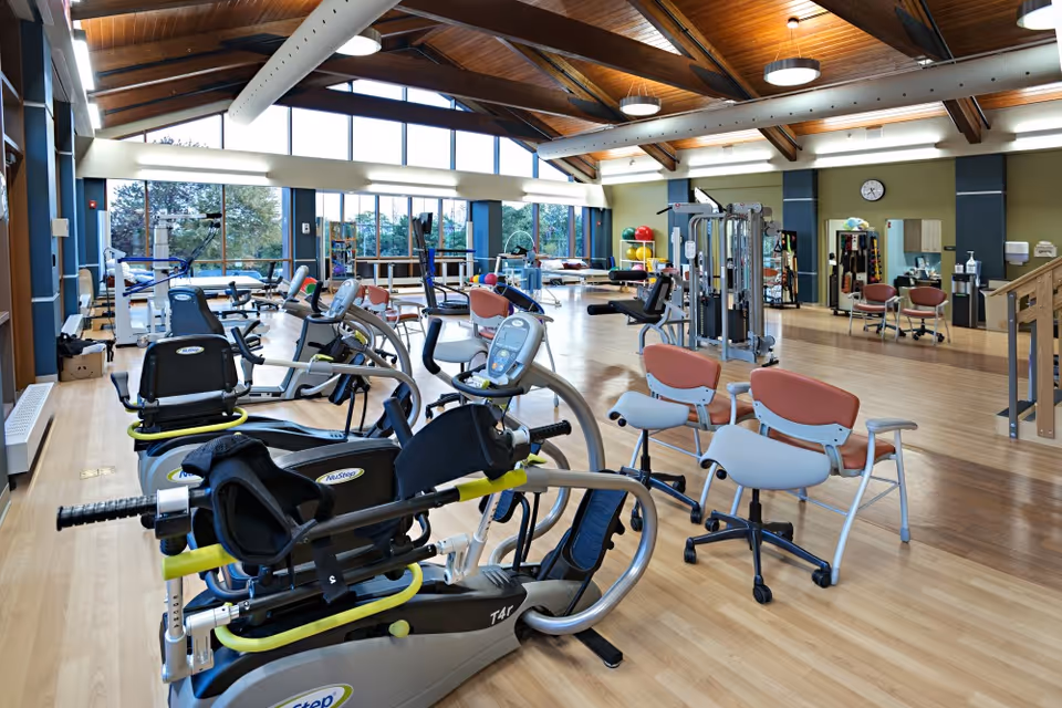 A spacious fitness room with various exercise equipment including recumbent bikes, weight machines, and exercise balls. The room has large windows allowing natural light to fill the space, wooden floors, and a high wooden ceiling with exposed beams. There are also chairs and mirrors along the walls.