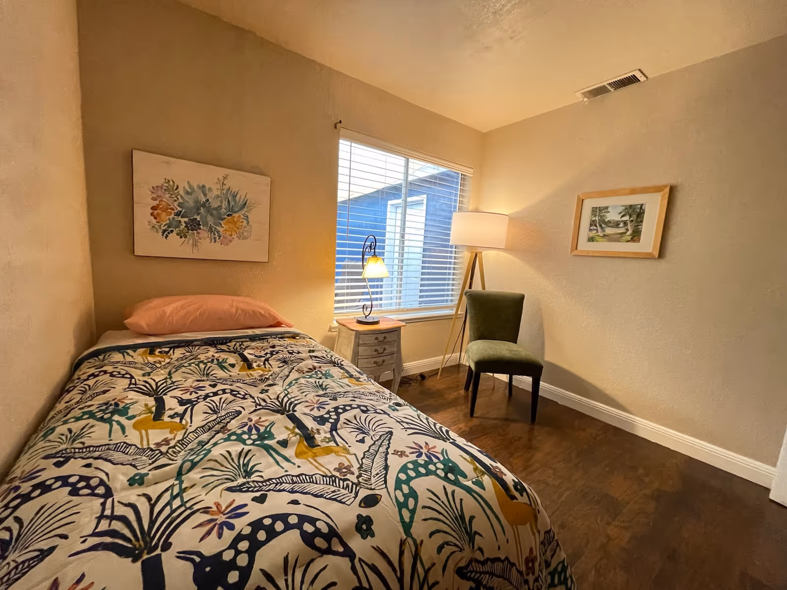Small furnished bedroom with a single bed, patterned bedcover, nightstand and lamp, a chair, and a window.