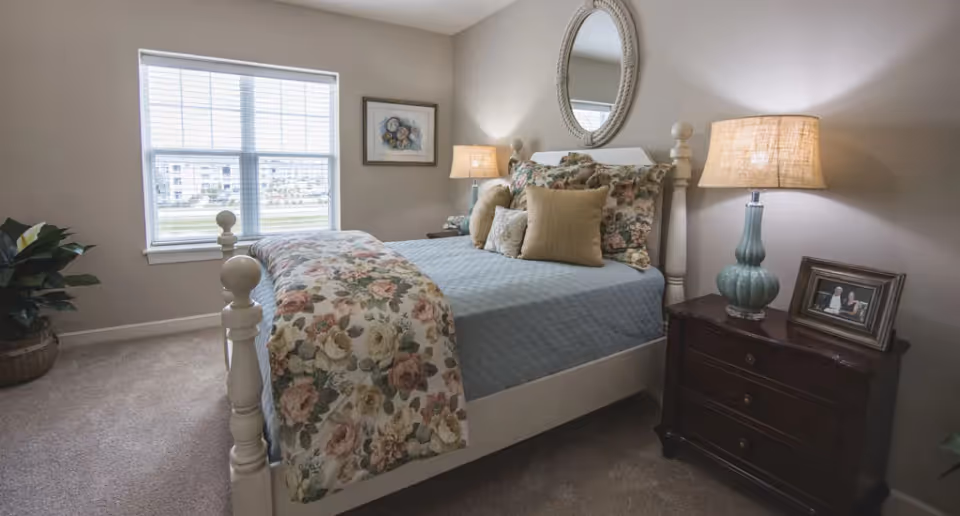 A cozy bedroom with a white wooden bed frame, a blue quilted bedspread, and floral-patterned pillows and blanket. There is a round mirror hanging above the bed, a nightstand with a lamp and a framed photo, a window with blinds letting in natural light, and a potted plant in the corner.