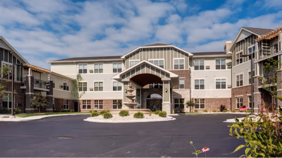 Front exterior of a multi-story senior living facility with a covered entrance, central fountain, and landscaped driveway under a partly cloudy sky.