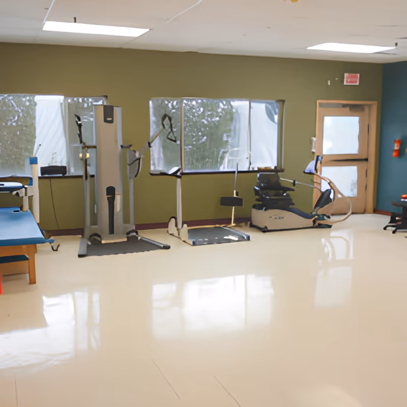 A bright rehabilitation/exercise room with medical exercise equipment, a recumbent bike, parallel bars and a treatment table near windows.