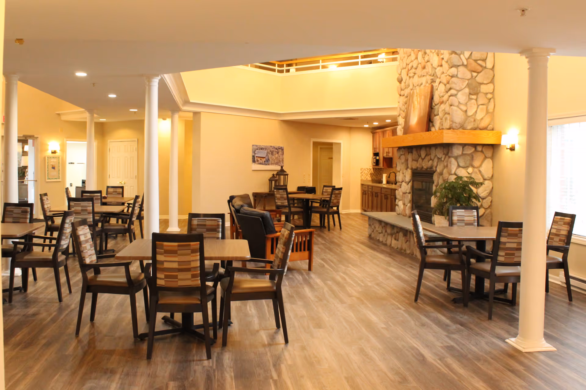 A spacious dining area with multiple wooden tables and chairs arranged neatly. The room features a stone fireplace with a wooden mantel, a small kitchenette area, and large windows allowing natural light to fill the space. The flooring is wood-style, and the walls are painted in warm neutral tones. White columns support the ceiling, adding an elegant touch to the room.