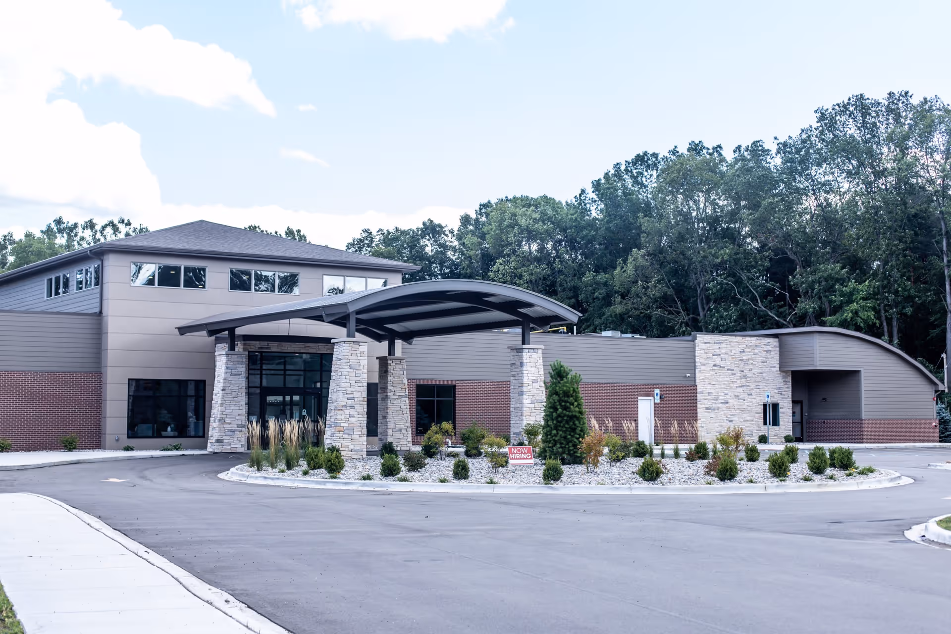 Exterior view of a modern senior living facility building with a covered entrance supported by stone pillars, surrounded by landscaped greenery and a circular driveway, with trees in the background under a partly cloudy sky.