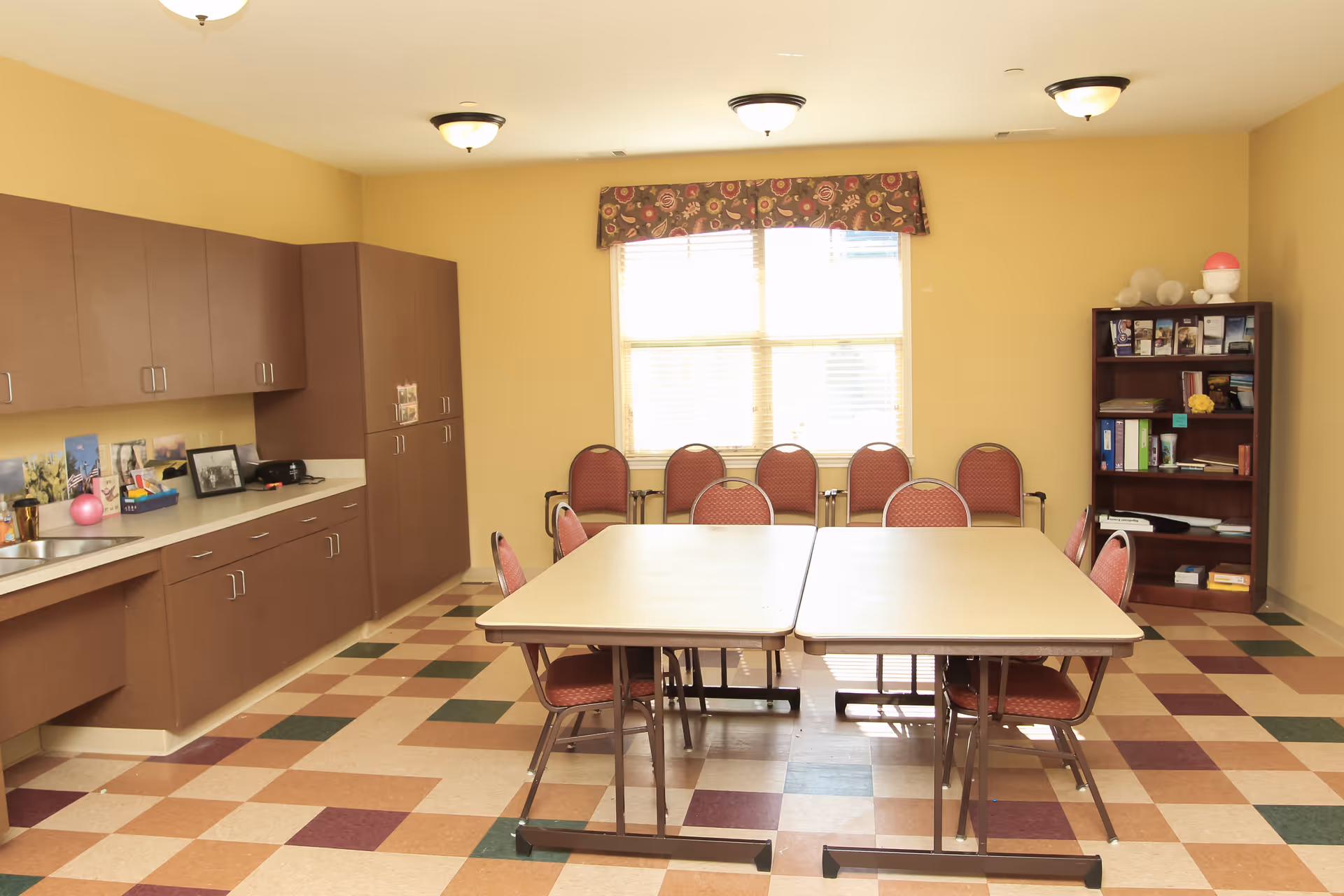 A well-lit room with beige walls and a checkered floor featuring shades of brown, green, and beige. There are two rectangular tables pushed together in the center, surrounded by red cushioned chairs. Along the left wall, there are brown cabinets with a countertop holding various items including framed photos and a pink ball. At the back of the room, a window with a floral valance lets in natural light. To the right, there is a wooden bookshelf with books and decorative items.