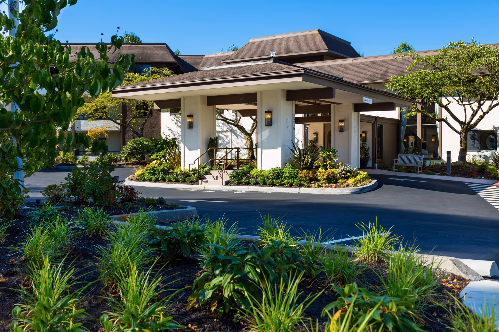 Entrance of Madison House Assisted Living Community with a covered drop-off area, surrounded by landscaped greenery and trees under a clear blue sky.