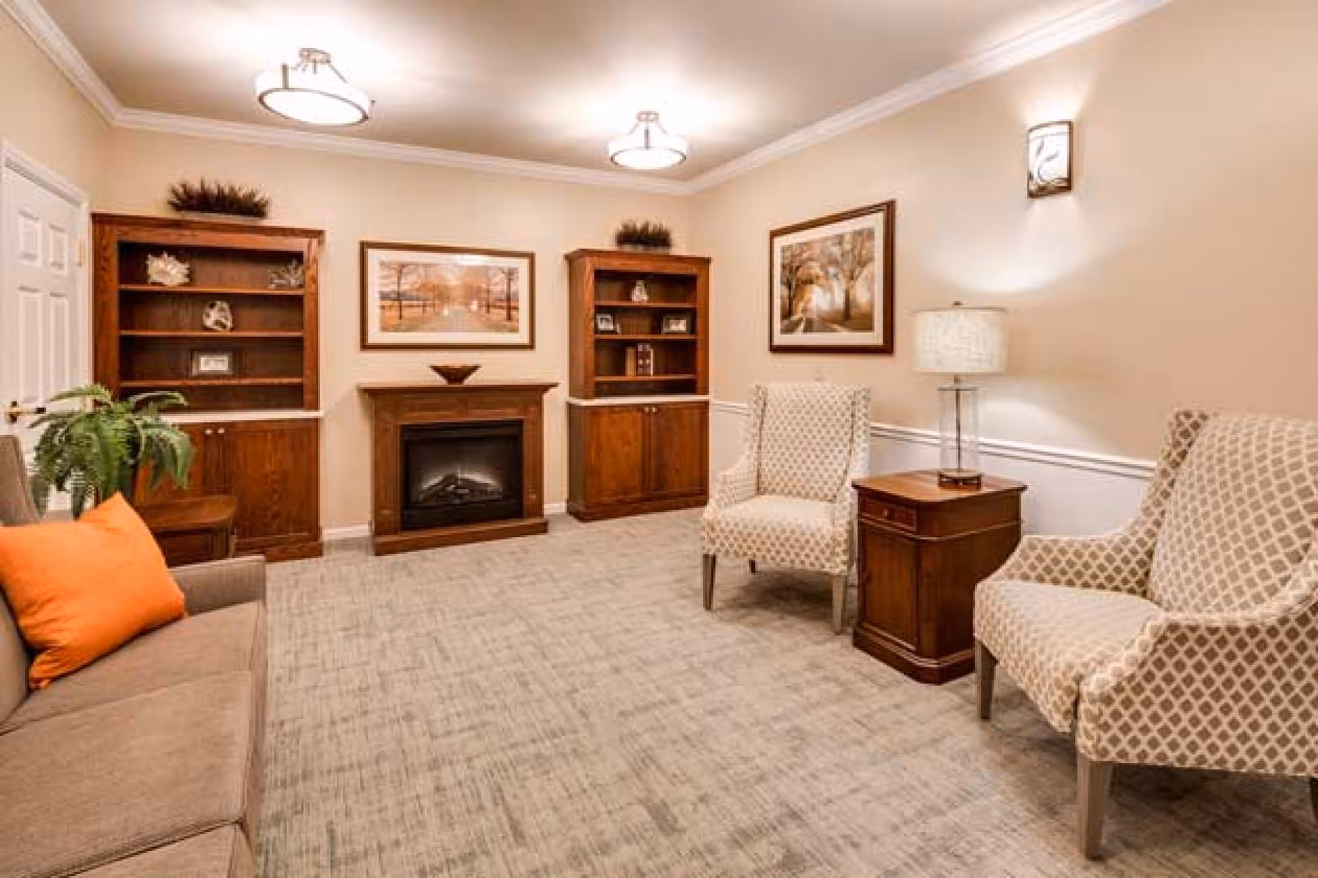 A cozy living room with beige walls and carpeted floor, featuring a brown sofa with an orange pillow on the left, two patterned armchairs on the right, a wooden side table with a lamp, two wooden bookshelves flanking a fireplace, and framed landscape artwork above the fireplace and on the wall.