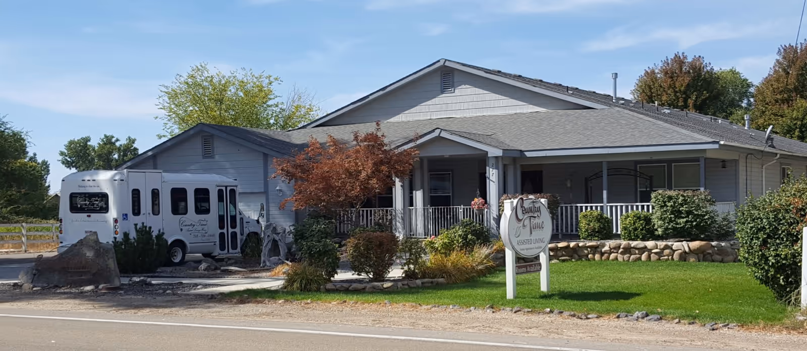 Exterior view of Country Time Assisted Living facility showing a single-story building with a porch, surrounded by greenery and trees. A white shuttle bus is parked near the building, and a sign with the facility's name is visible on the lawn.
