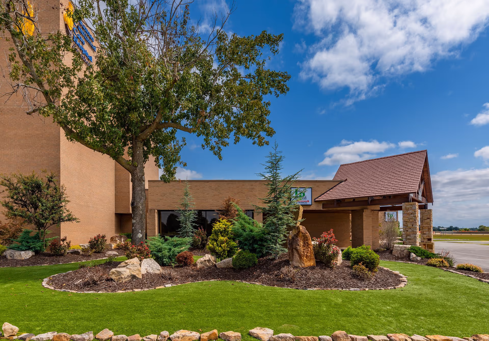 Front exterior of an assisted living building with a landscaped lawn, trees, rocks, and a covered entrance under a blue sky.