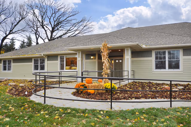 Exterior view of a single-story building with light green siding and a gray roof. The entrance has a ramp with black railings and a small landscaped area featuring pumpkins, flowers, and a bundle of dried corn stalks. Trees and a partly cloudy sky are visible in the background.