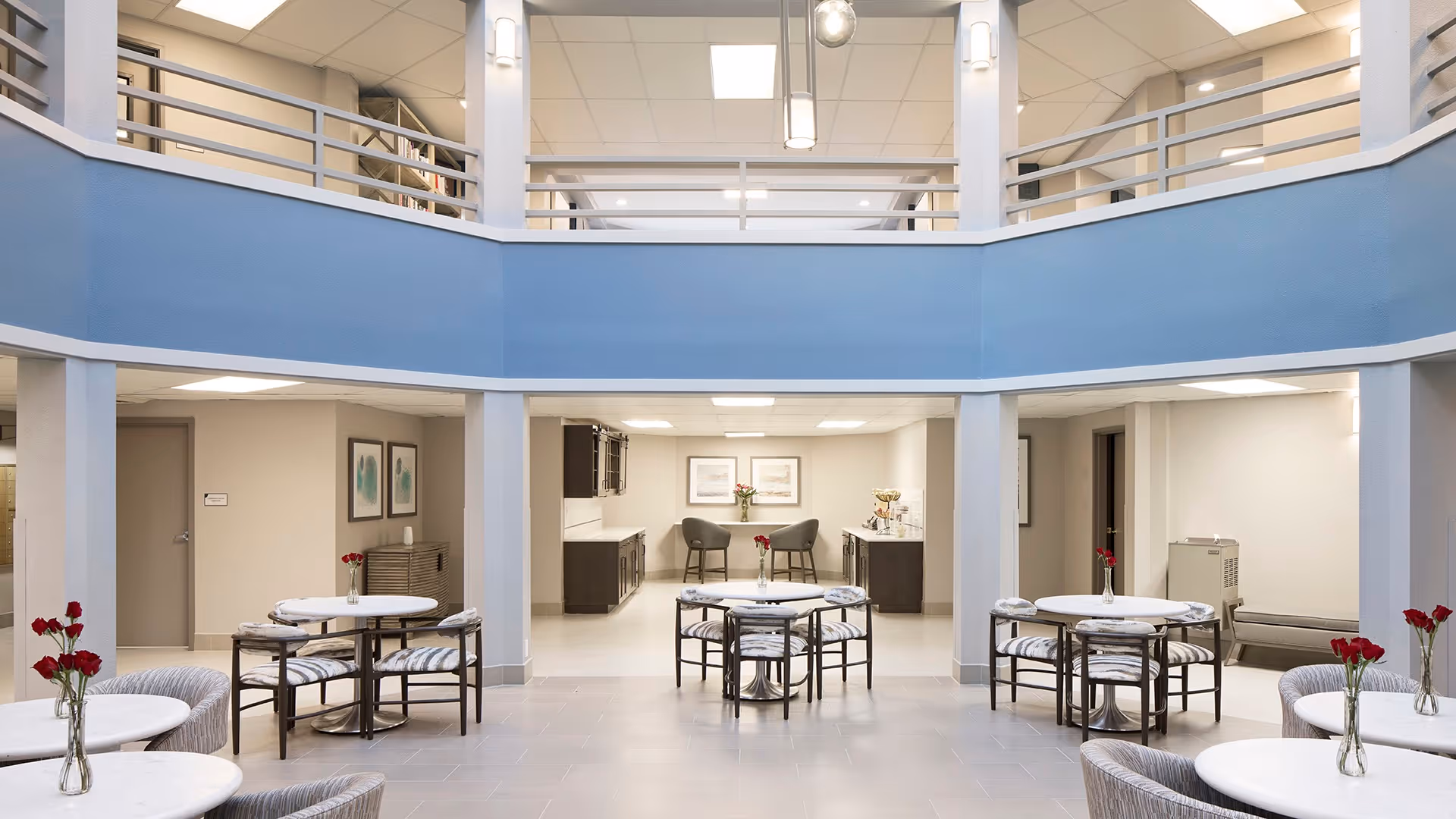 A bright, open interior common area with a high ceiling and a second-floor balcony railing. The space features several round tables with chairs arranged around them, each table decorated with a small vase of red flowers. In the background, there is a seating area with two chairs and framed artwork on the wall, along with cabinetry and a countertop. The walls are painted in neutral tones with a blue accent band around the upper level.