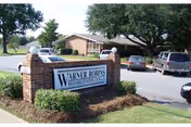Exterior view of Warner Robins Rehabilitation Center showing a brick sign with the facility name in front of a parking lot with several parked cars and a single-story building surrounded by trees and greenery.