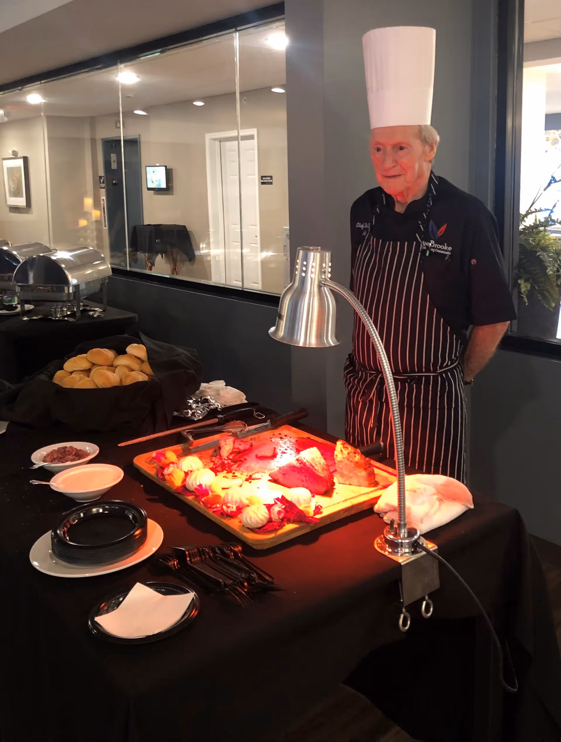 An elderly chef wearing a tall white chef hat and striped apron stands behind a buffet table with a carving station. The table has a heat lamp shining on sliced meat and vegetables on a wooden cutting board, with plates, utensils, rolls, and sauces nearby. The background shows a glass wall and a hallway.