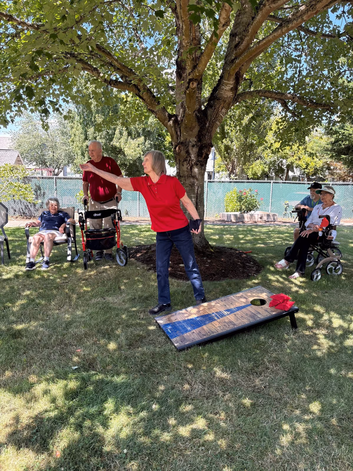 A group of elderly people enjoying an outdoor game of cornhole under a large tree. One woman in a red shirt is actively throwing a bean bag towards the cornhole board on the grass, while others are seated nearby watching and relaxing in the shade.