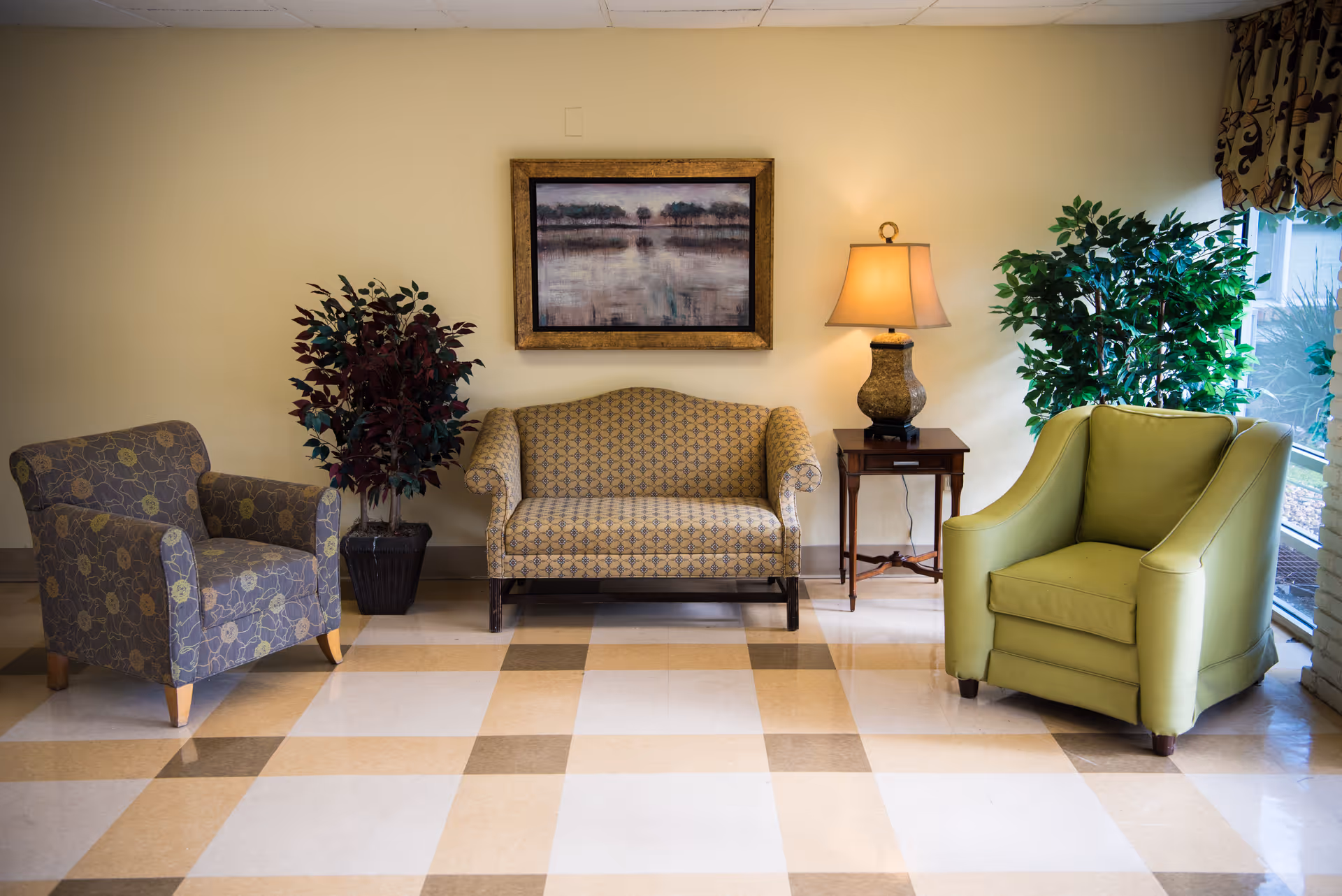 A sitting area with two armchairs and a patterned loveseat flanked by plants, a side table with a lamp, and framed artwork on the wall.