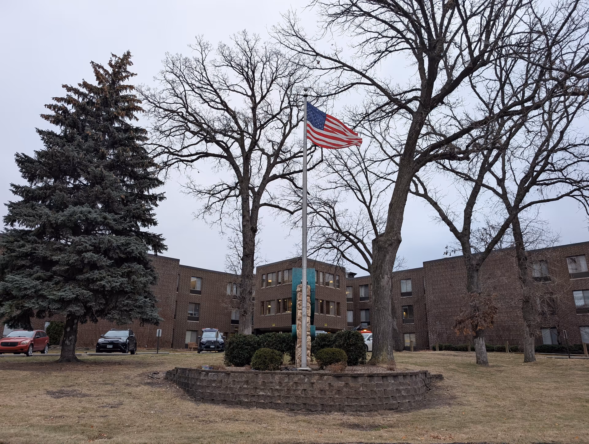 Brick building front with an American flag on a flagpole, leafless trees, and a grassy lawn.