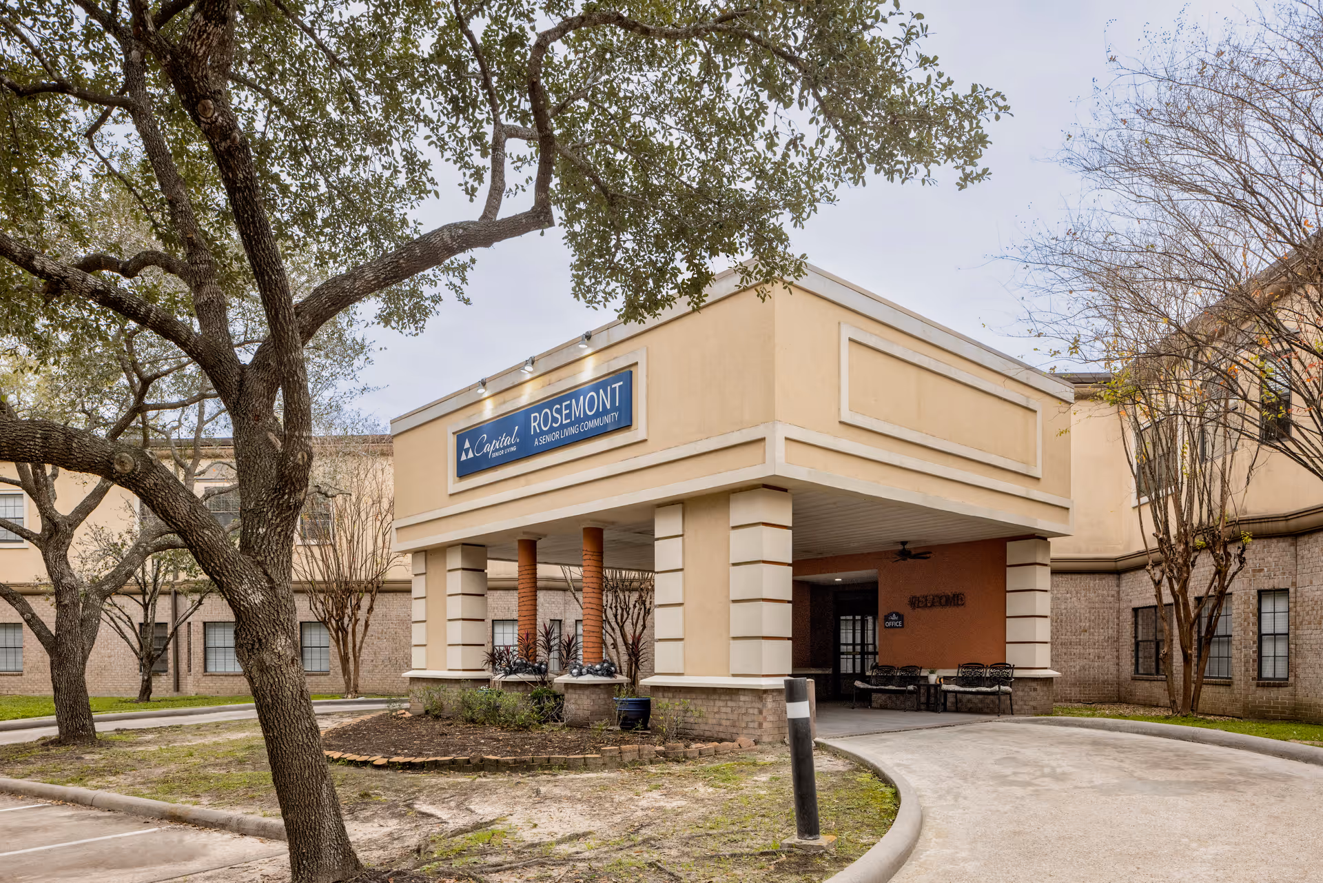 Exterior view of the entrance to Rosemont Assisted Living And Memory Care facility, showing a covered drop-off area supported by columns, surrounded by trees and a curved driveway.