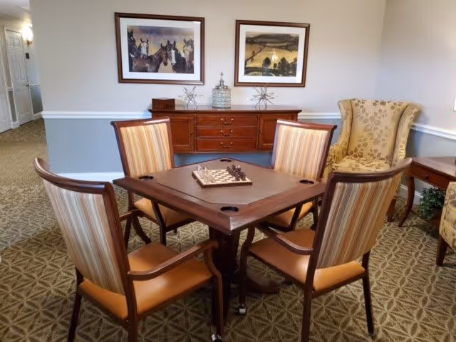 A cozy common area with a square wooden table set for chess, surrounded by four striped cushioned chairs. Behind the table is a wooden sideboard with decorative items and two framed paintings of horses and a landscape on the wall. To the right, there is a patterned armchair and a small side table with a plant underneath. The room has carpeted flooring and soft lighting.