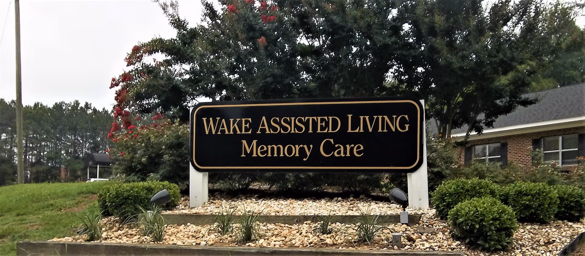 Outdoor view of a landscaped area with a black and gold sign that reads 'WAKE ASSISTED LIVING Memory Care' surrounded by bushes, small plants, and trees with a building partially visible in the background.