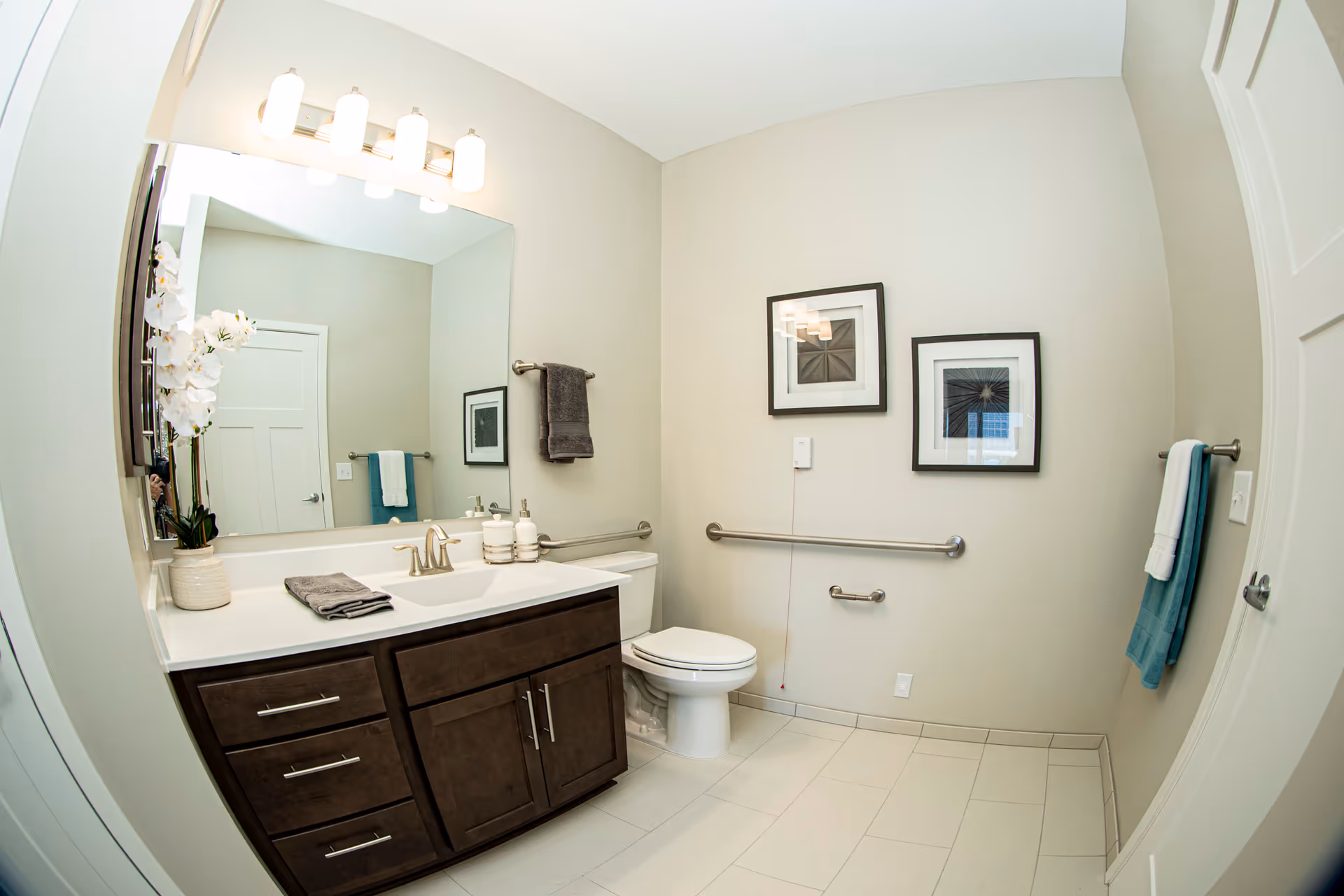 A clean, modern bathroom with a large mirror above a dark wood vanity with a white countertop. The vanity has a sink with a silver faucet, a gray hand towel, and decorative items including a white orchid plant. There is a toilet next to the vanity with grab bars on the wall for accessibility. Two framed pictures hang on the wall above the toilet. The bathroom has light-colored walls and floor tiles, and there are two towel racks with towels on either side of the room.