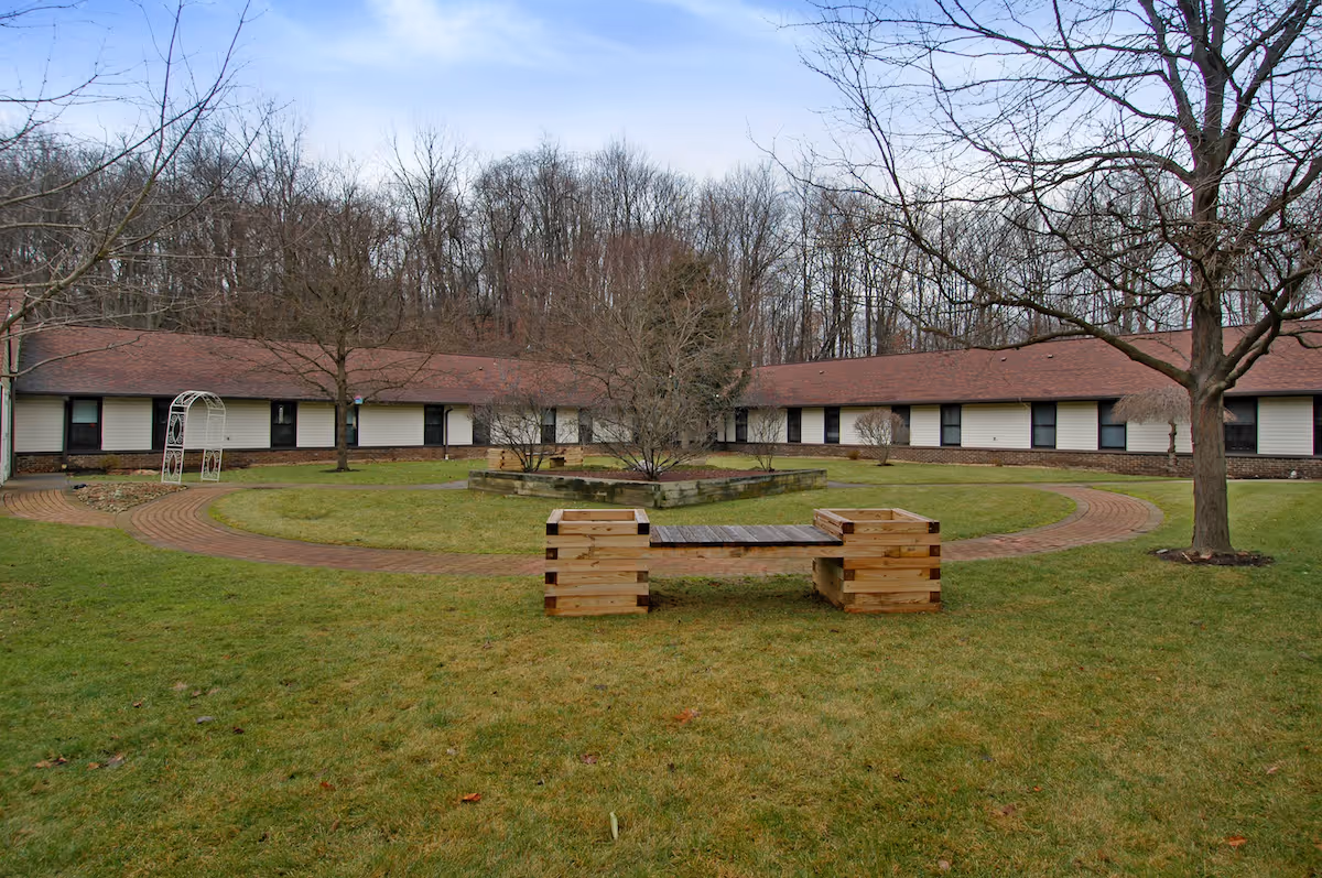 Courtyard with a circular brick walkway, grassy lawn, wooden bench, bare trees, and a surrounding single-story residential building.