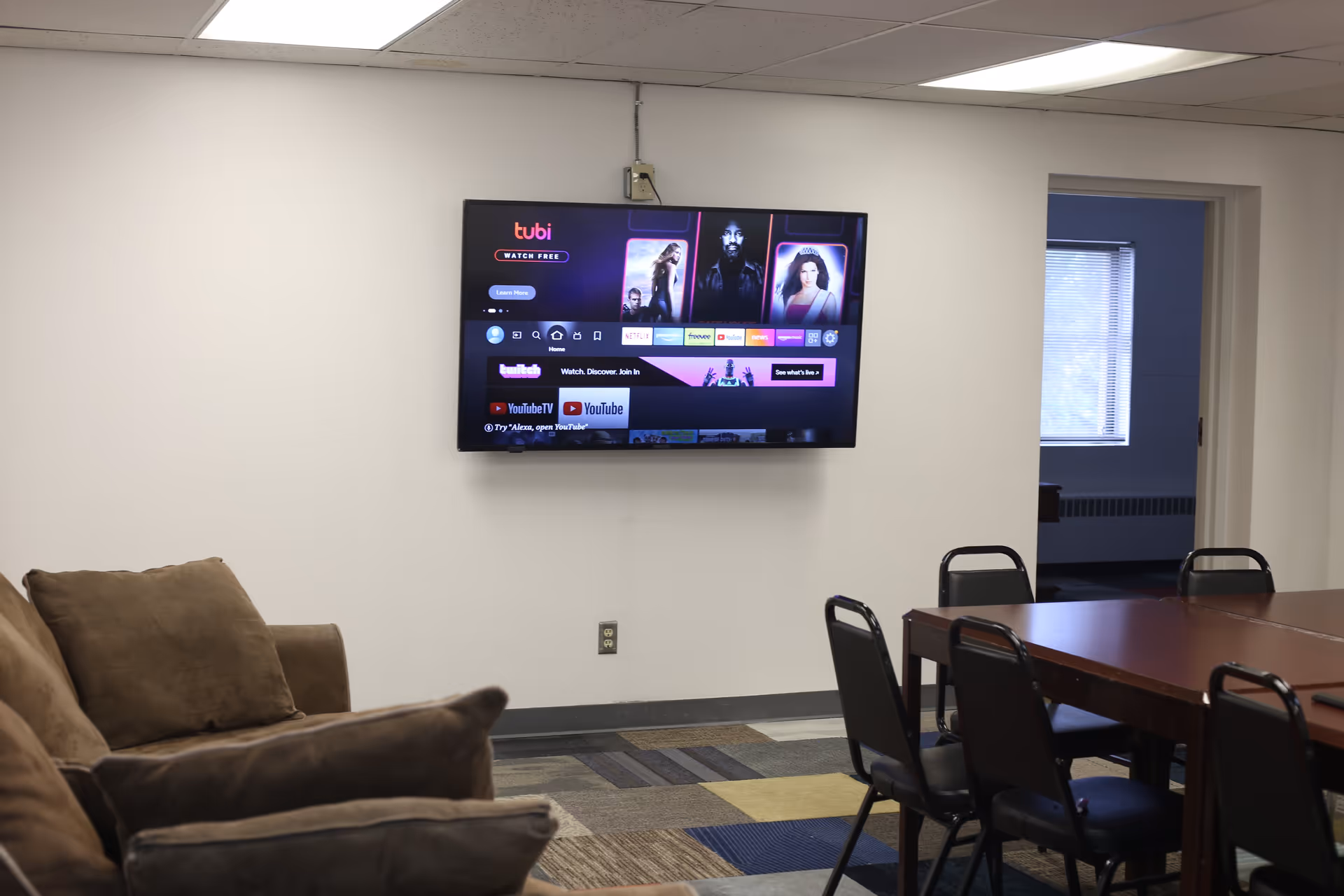 Wall-mounted TV in a community room with sofas on the left and a table with chairs on the right.
