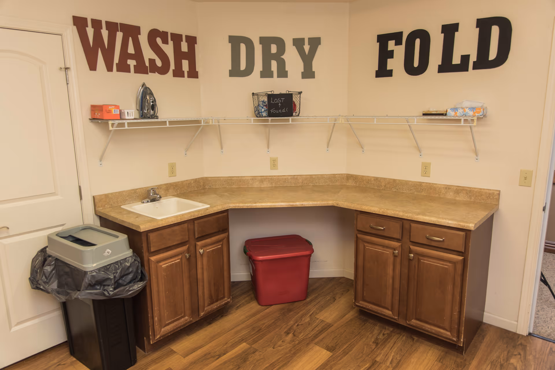 Corner laundry room with countertop and sink, wooden cabinets, trash and storage bins, and large wall letters reading 'WASH DRY FOLD'.