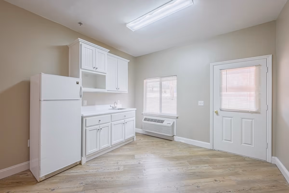 A small kitchen area with white cabinets, a white refrigerator, a sink, a window with blinds, a door with a window and blinds, and a wall-mounted air conditioning unit below the window. The floor is wooden and the walls are painted light beige.