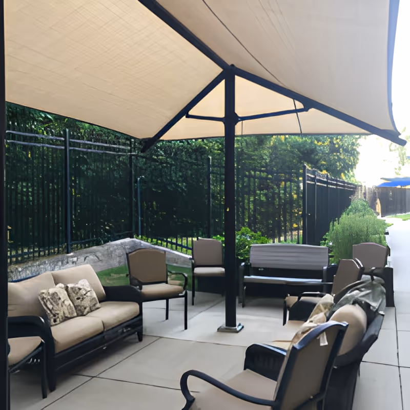 Shaded outdoor patio with cushioned chairs and sofas arranged around a central umbrella on a paved terrace next to a fence and greenery.