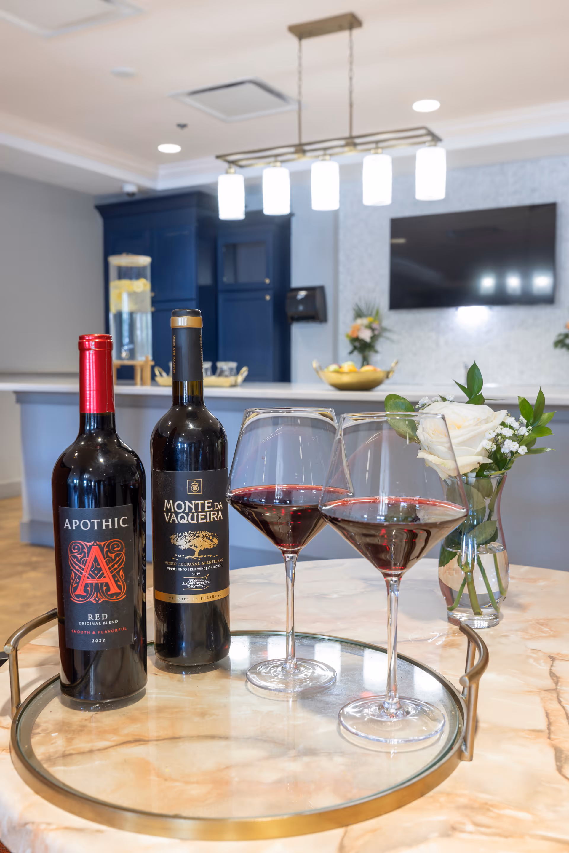 Two bottles of red wine and two glasses filled with red wine on a round tray placed on a marble table. In the background, there is a modern kitchen area with blue cabinets, a countertop with a bowl of fruit, a water dispenser, a wall-mounted TV, and a vase with white flowers.