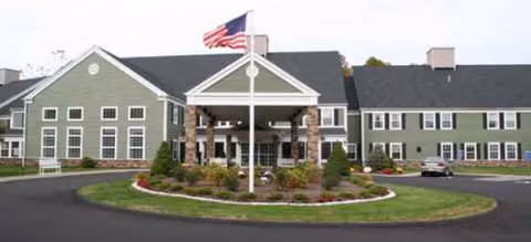 Green, two-story senior living facility with a central entrance, flagpole, and circular landscaped driveway.