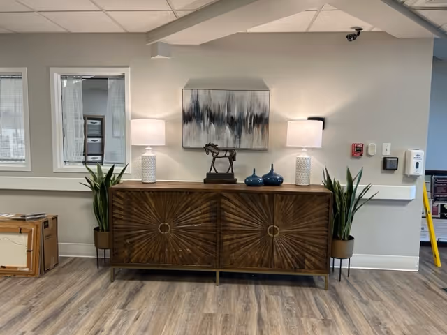 A wooden sideboard with a sunburst pattern on its doors is placed against a light gray wall. On top of the sideboard are two white table lamps with textured bases, a small horse sculpture, and two blue decorative vases. Two tall potted plants flank the sideboard. Above it hangs an abstract painting with shades of gray, black, and white. The floor is wood laminate, and there are two windows with blinds on the left side of the wall.