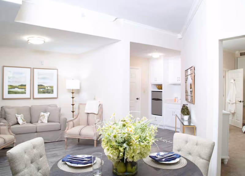 A bright and airy living and dining area in a senior living facility. The space features a round dining table set with plates, napkins, and glasses, and a centerpiece of green and white flowers. In the background, there is a beige sofa with patterned pillows, two upholstered armchairs, a floor lamp, and framed landscape artwork on the wall. The room has light-colored walls, wood flooring, and an open doorway leading to a kitchen area with white cabinetry and appliances.