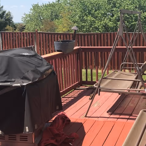 Red wooden deck with grill, swing, and potted plant overlooking trees.