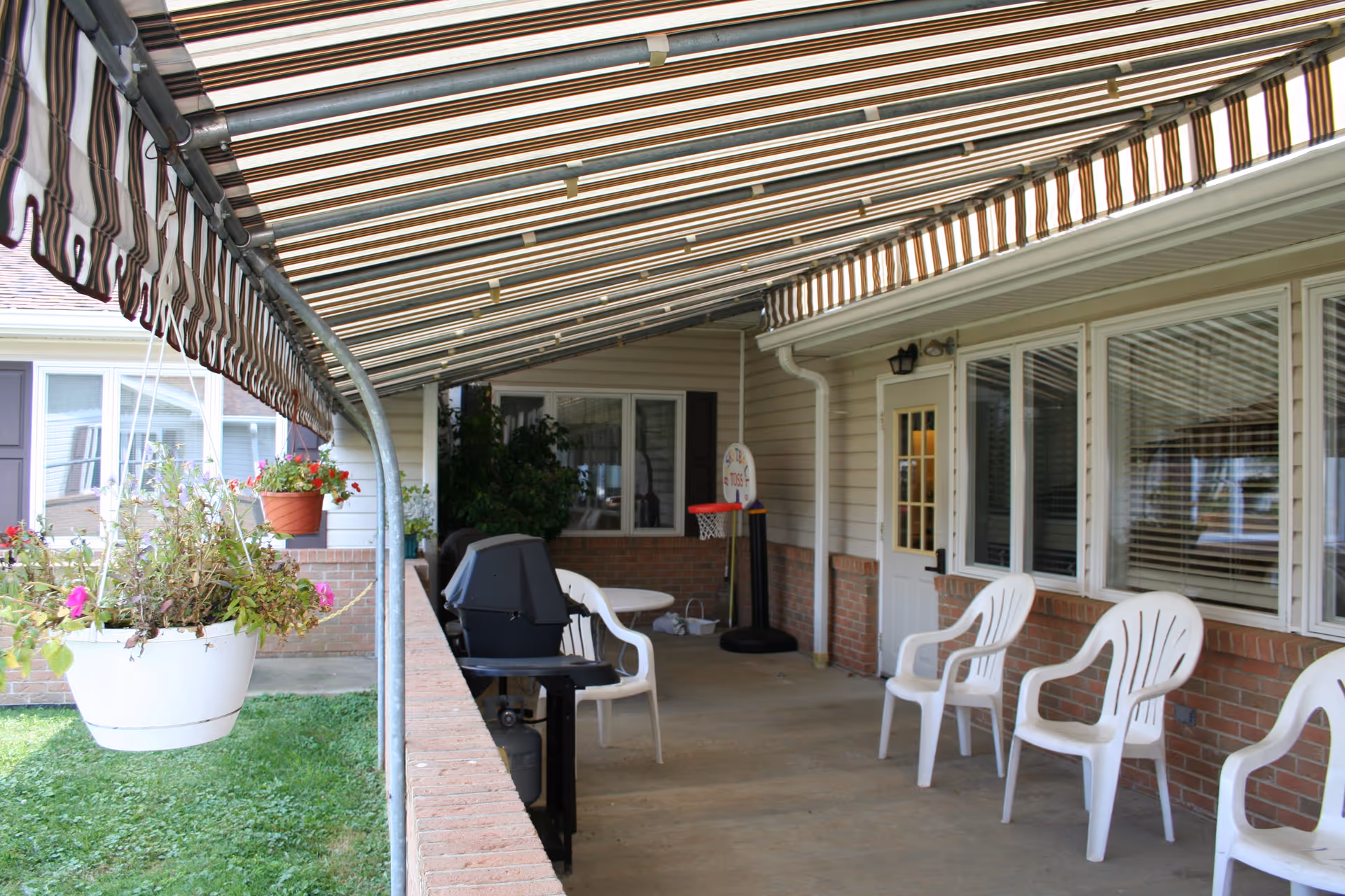 Covered outdoor patio area with a striped awning, white plastic chairs, a small grill, hanging flower pots, and a small basketball hoop near a door and windows.