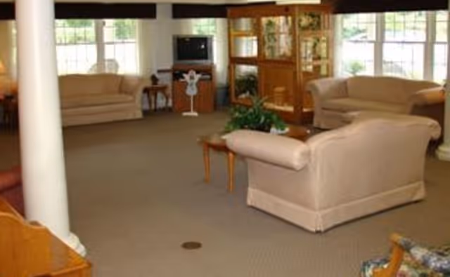 A spacious living room with beige sofas arranged around a small wooden coffee table with a plant on it. There is a wooden display cabinet and a TV on a stand against the far wall, with large windows letting in natural light.